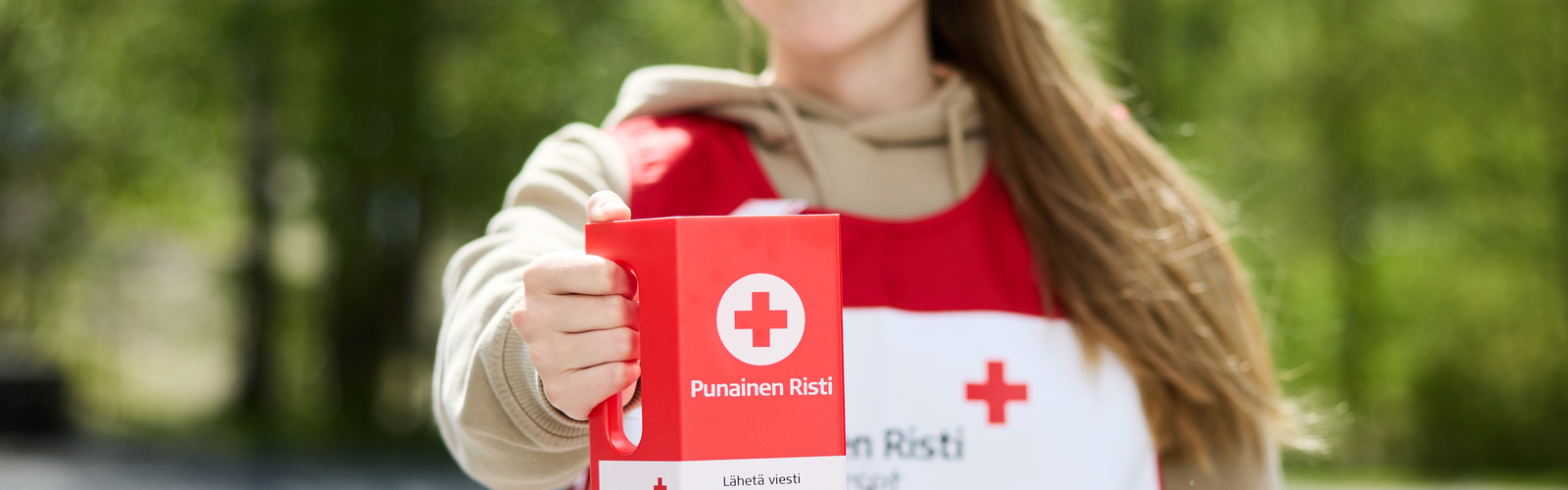 Hymyilevä henkilö Punaisen Ristin liivissä keräyslipas kädessään.
